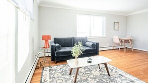A living room with a dark blue sofa, a white coffee table, and a small side table with a lamp. There is a dining area with a table and chairs.