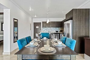 A modern dining area with a dark wooden table set for six, blue upholstered chairs, and a kitchen in the background with a tiled backsplash. - 2BR Condo w/ Pool & Gym, near Palm Beach (Noord)