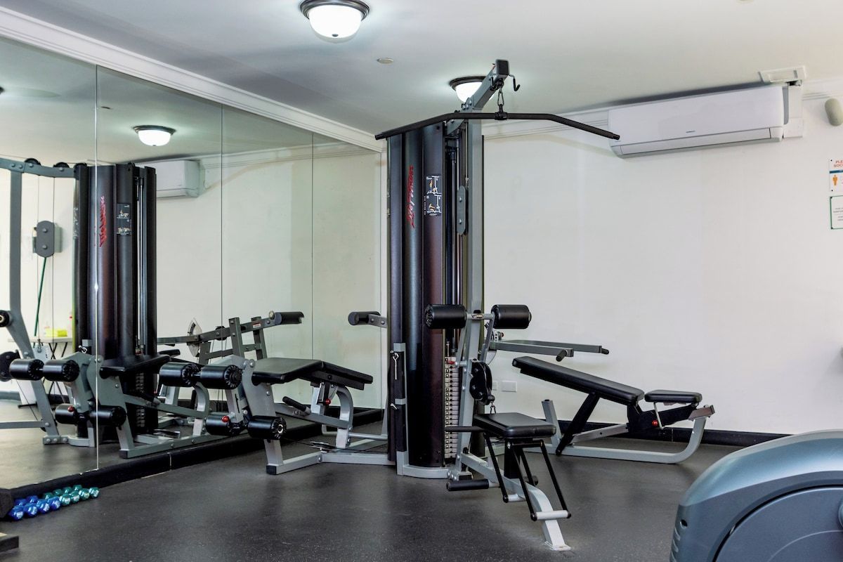 Un gimnasio con máquinas de pesas, una prensa de banca y un muro con espejos.