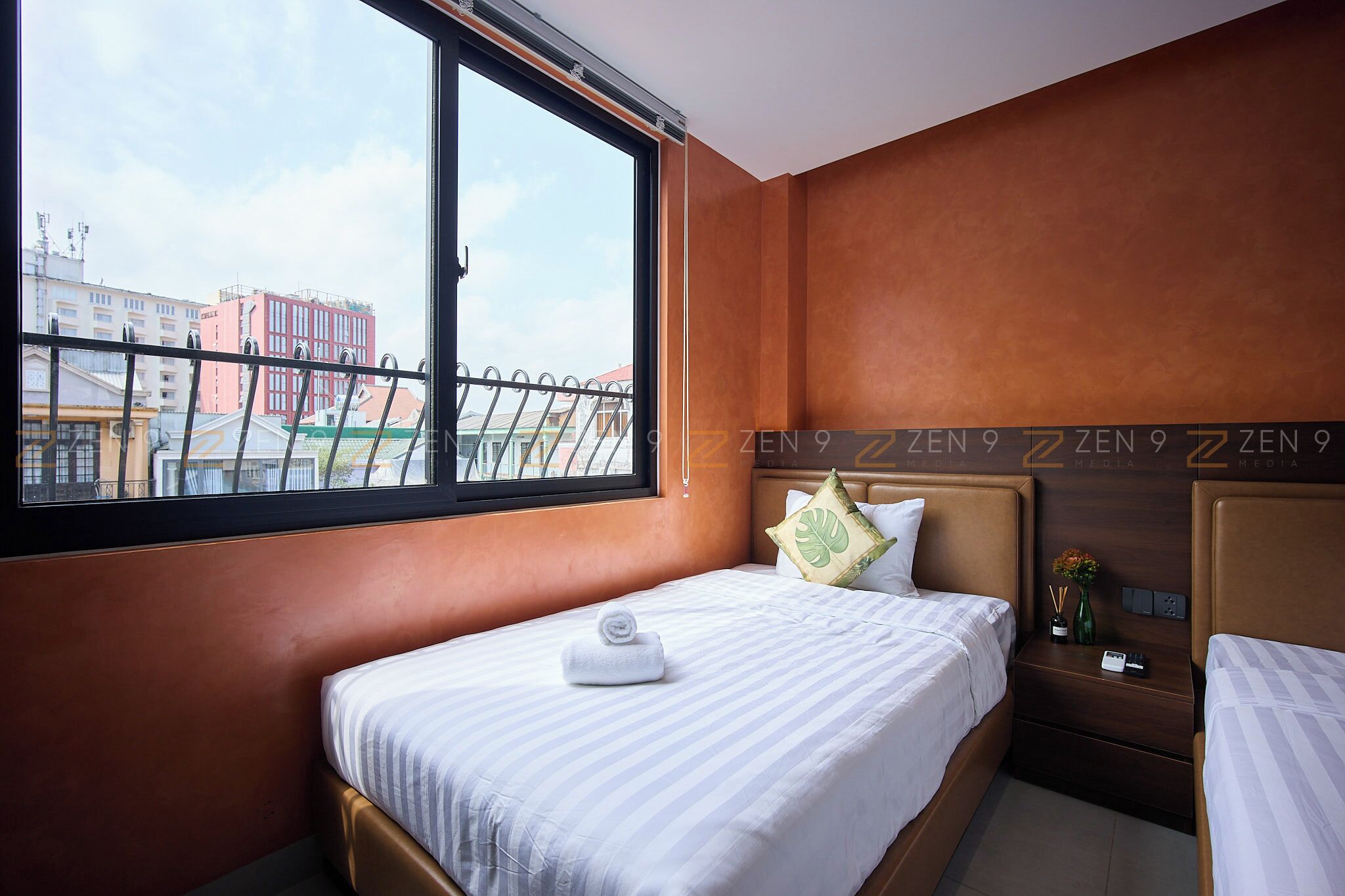 A hotel room with a large window, a bed with a rolled towel, a nightstand with a vase, and a headboard with the text "ZEN 9".