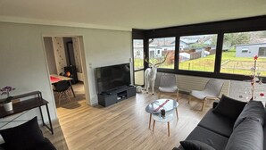 Un salon moderne avec une grande fenêtre, un canapé noir, une table basse en verre et un téléviseur.