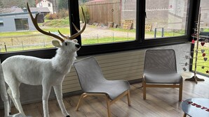 Une pièce dotée d’une grande fenêtre, d’une sculpture représentant la tête d’un cerf blanc, de deux fauteuils gris et d’une table ornée d’un élément décoratif portant l’inscription « JOY ».