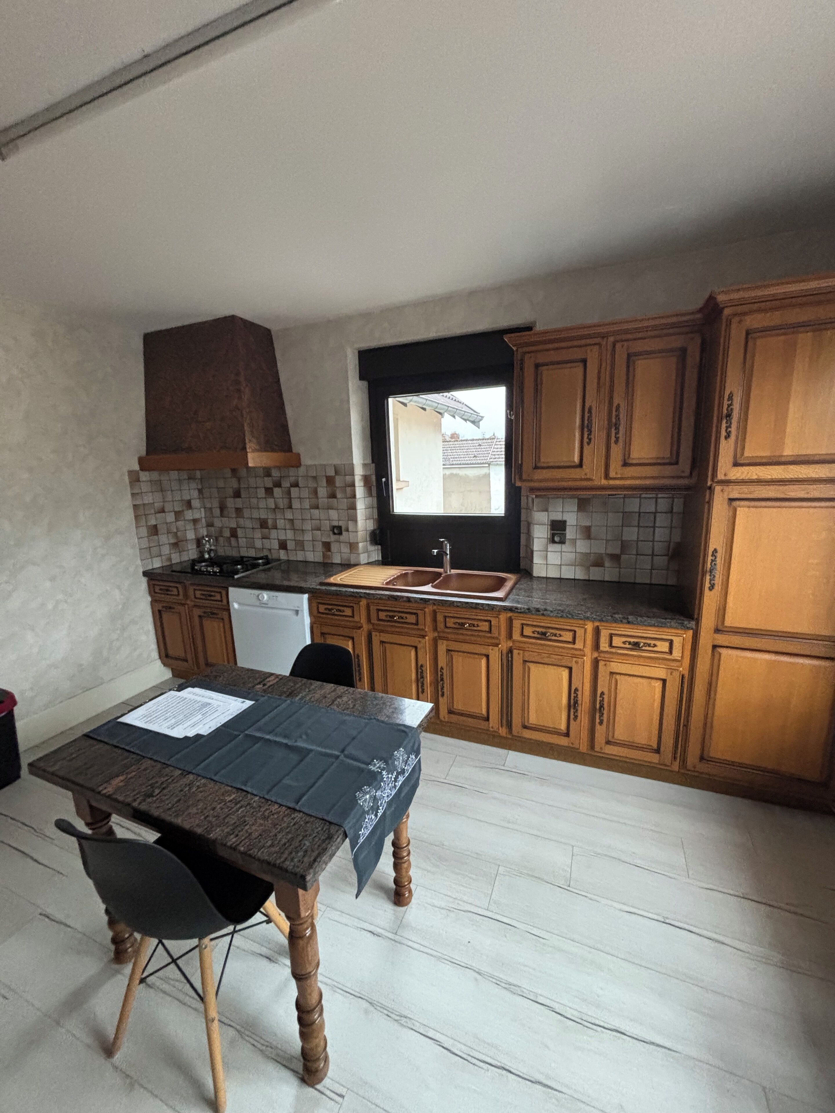 Una cucina compatta con mobili in legno, zoccolo in piastrelle, una finestra e un tavolo da pranzo con tovaglietta.