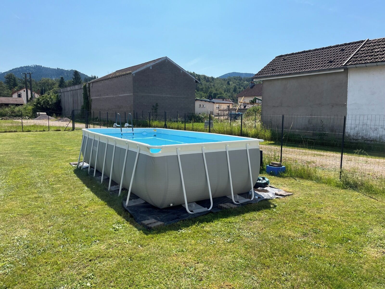 An above-ground swimming pool in a grassy yard with a metal frame, surrounded by a fence and houses in the background.