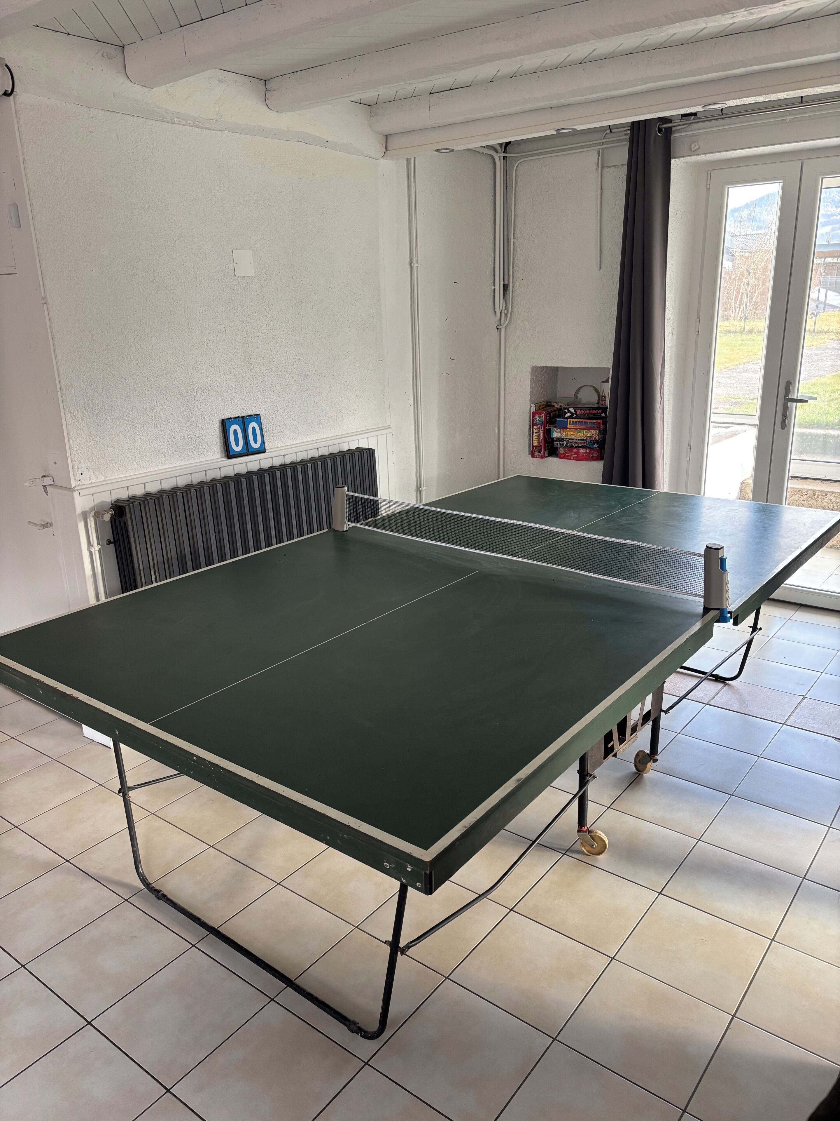Une table de ping-pong dans une pièce aux murs blancs, avec un radiateur et une porte donnant sur l’extérieur.