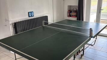 Une table de ping-pong dans une pièce aux murs blancs, avec un radiateur et une porte donnant sur l’extérieur.
