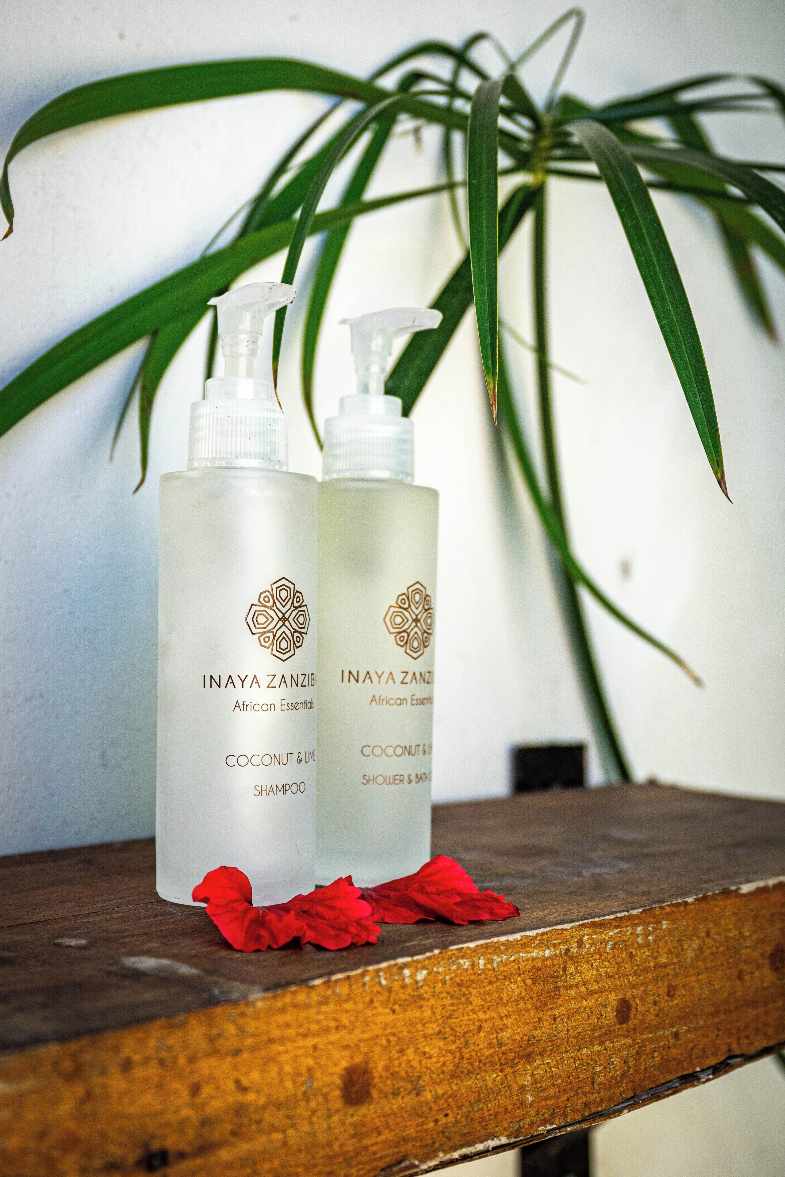 Dos botellas de champú Inaya Zanzibar African Essentials de coco y lima sobre una superficie de madera, con un pétalo de flor roja y una planta verde al fondo.