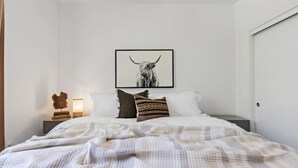 A bedroom with a bed, pillows, a framed picture, a nightstand, and a wall-mounted lamp. - Gardenia at Evergreen | A Stylish Flat In Downtown RDD (Redding)