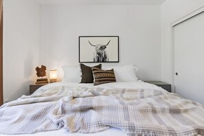 A bedroom with a bed, pillows, a framed picture, a nightstand, and a wall-mounted lamp.