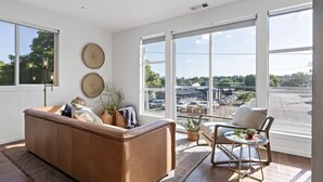A modern living room with a brown sofa, a small round table, and a chair. There are large windows with a view of a parking lot and trees. - Gardenia at Evergreen | A Stylish Flat In Downtown RDD (Redding)