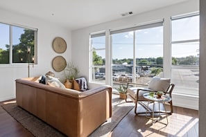 A modern living room with a brown sofa, a small round table, and a chair. There are large windows with a view of a parking lot and trees.