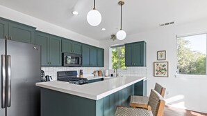 A modern kitchen with teal cabinets, a white island, stainless steel appliances, and a dining area with wicker chairs. - Gardenia at Evergreen | A Stylish Flat In Downtown RDD (Redding)