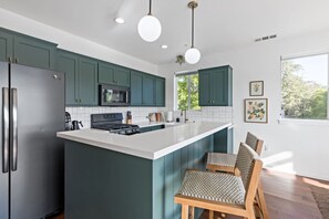 A modern kitchen with teal cabinets, a white island, stainless steel appliances, and a dining area with wicker chairs.