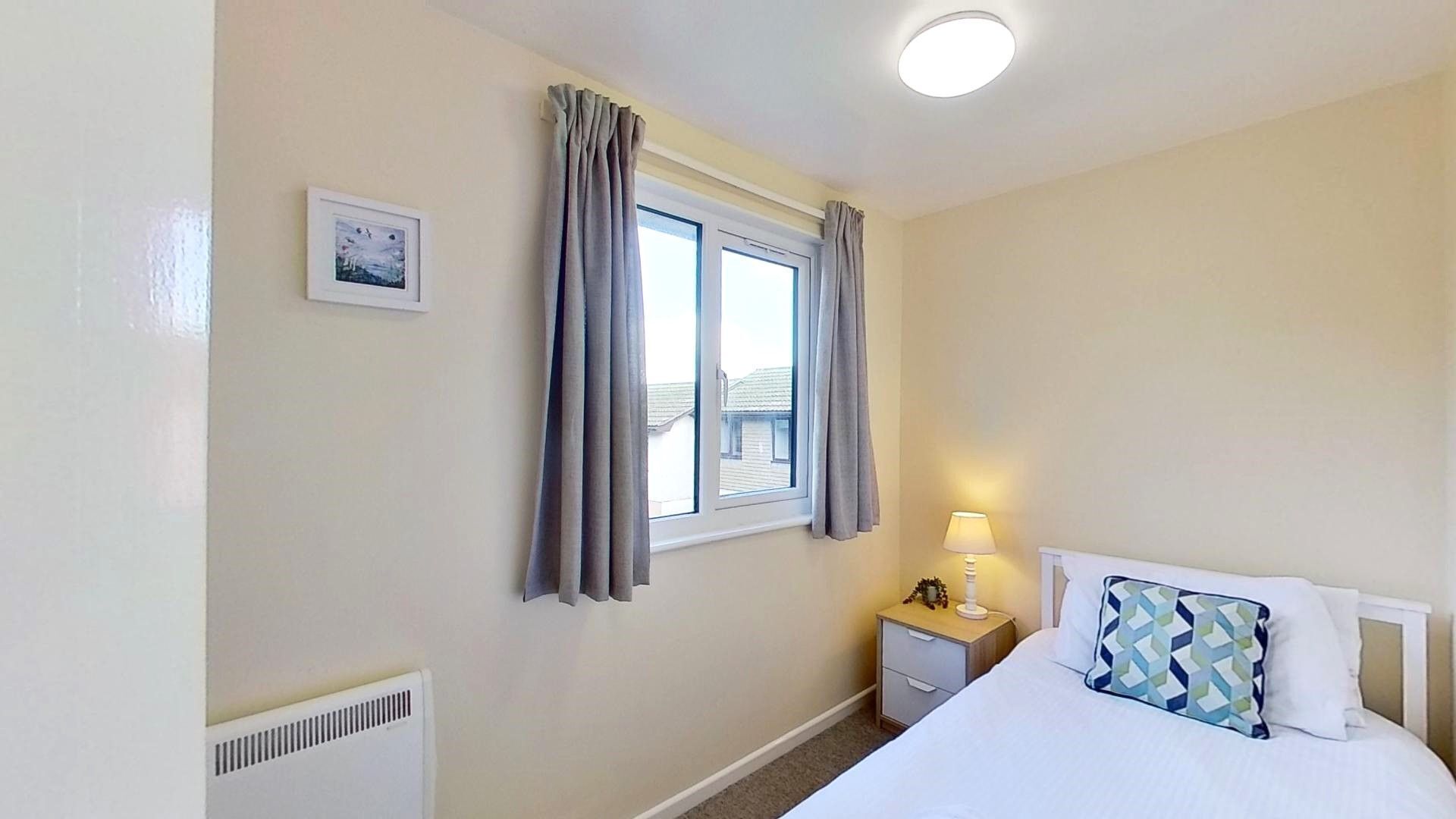 Een slaapkamer met een bed, een nachtkastje met een lamp, een raam met gordijnen en een radiator.