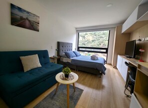 Een moderne kamer met een bank, een bed, een kleintje tafeltje met een plant, een televisie en een groot raam met uitzicht op groen.