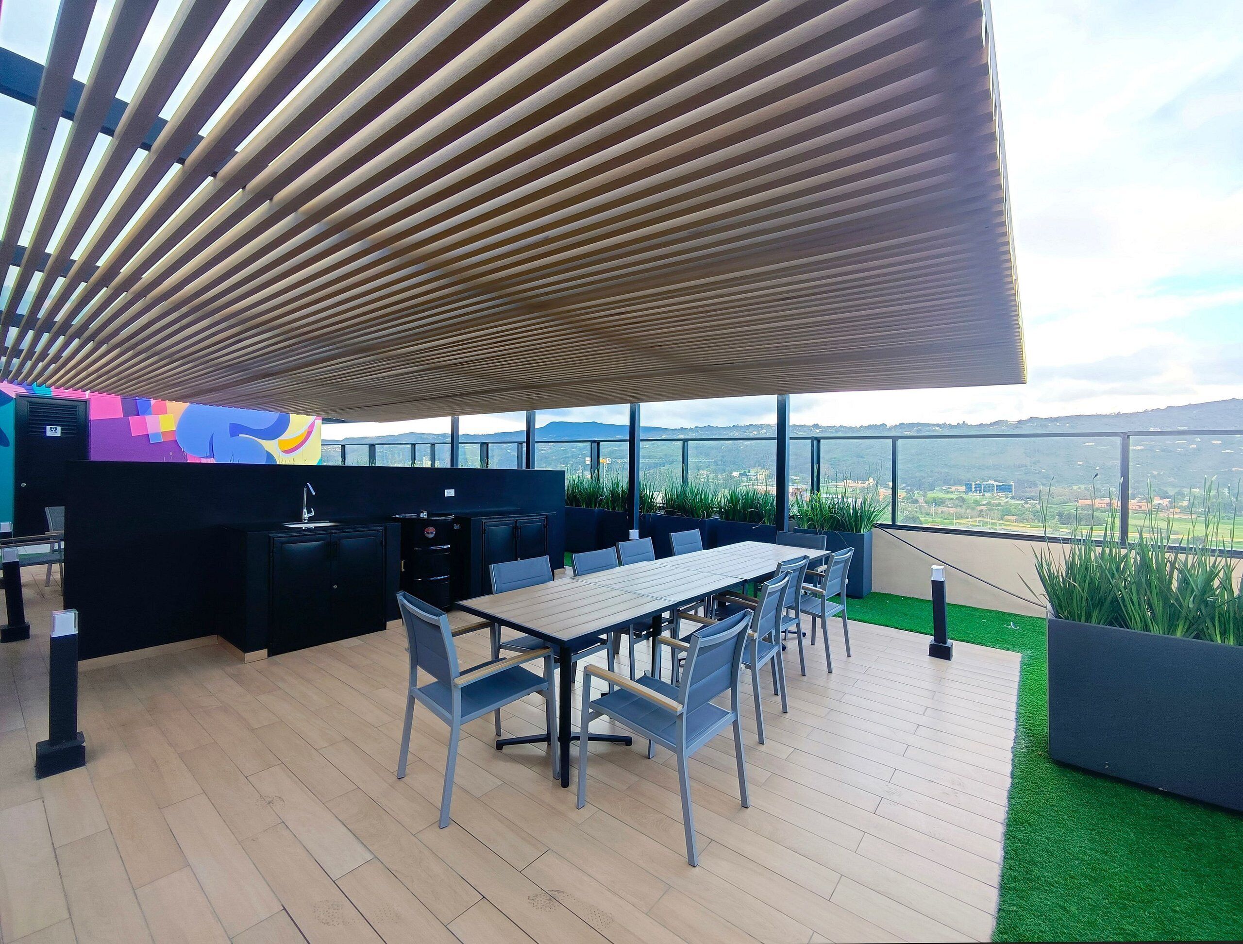 Un espace de restauration extérieur moderne, doté d’un plafond en bois, d’une longue table et de chaises, offrant une vue imprenable sur un paysage pittoresque.