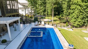 Une piscine rectangulaire avec un petit jacuzzi au centre, entourée d’un patio aménagé avec du mobilier d’extérieur et un espace salon couvert.