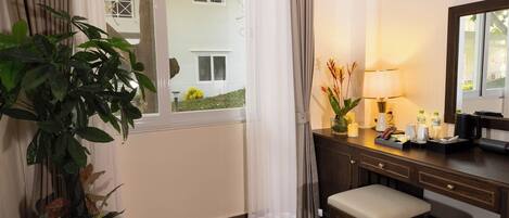 A hotel room with a wooden desk, a bench, a mirror, a window with curtains, and a potted plant.