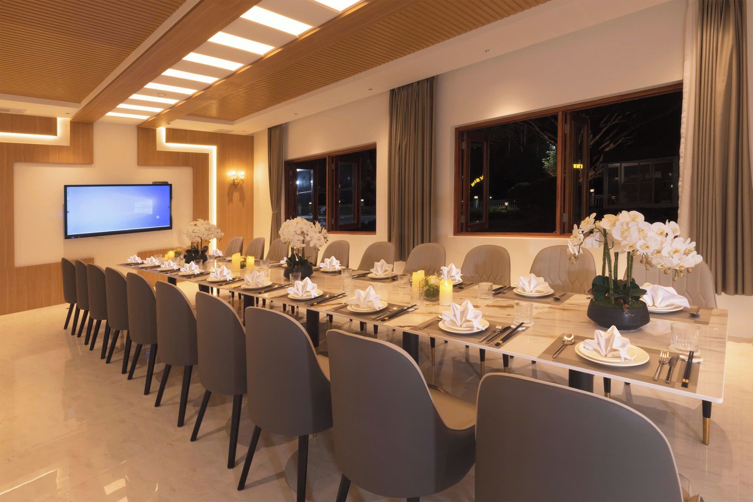 Un salón de conferencias con una larga mesa dispuesta para una reuni ón, equipado con sillas, un televisor de pantalla plana y elementos decorativos como flores y velas.