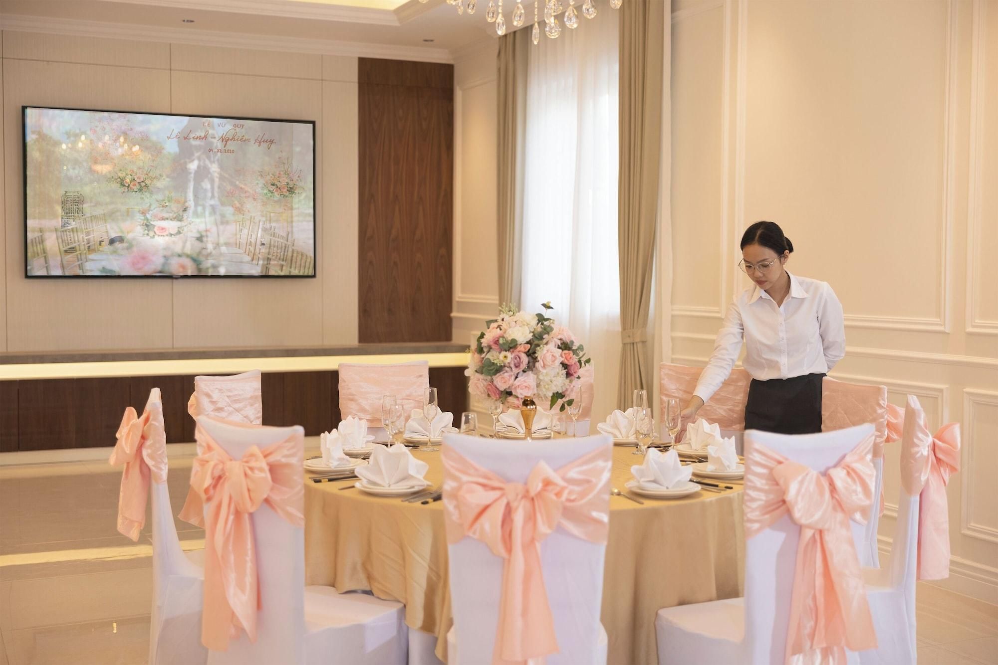 Un salón de banquetes con mesas dispuestas para un evento formal, un gran centro de flores y un miembro del personal atendiendo la mesa.