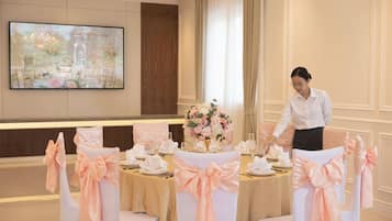 Un salón de banquetes con mesas dispuestas para un evento formal, un gran centro de flores y un miembro del personal atendiendo la mesa.