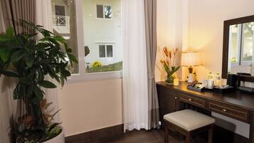 A hotel room with a wooden desk, a chair, a potted plant, a lamp, and a window with curtains.