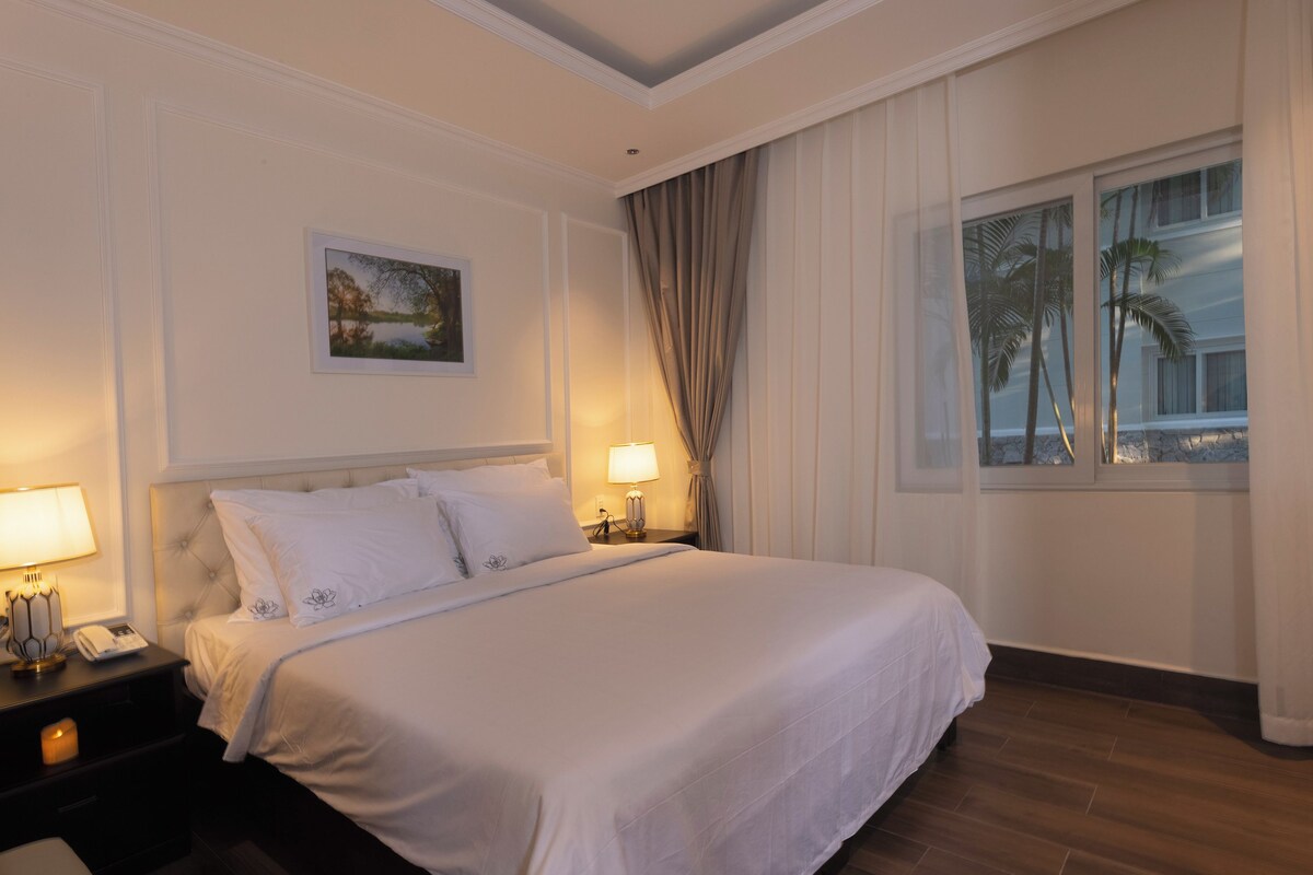 Quarto de hotel com uma cama grande, uma mesa de cabeceira com abajur, um quadro na parede e uma janela com cortinas.
