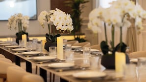 Una larga mesa de comedor dispuesta con platos blancos, vasos transparentes y velas, rodeada de sillas.