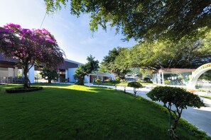 A well-maintained garden with a variety of trees and shrubs, a paved walkway, and a building with a modern architectural style. - Merperle Núi Sam Resort (Vinh Te)