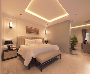A modern hotel bedroom with a large bed, bedside tables, a bench, a potted plant, and a view of the adjacent living area. - Merperle Núi Sam Resort (Vinh Te)