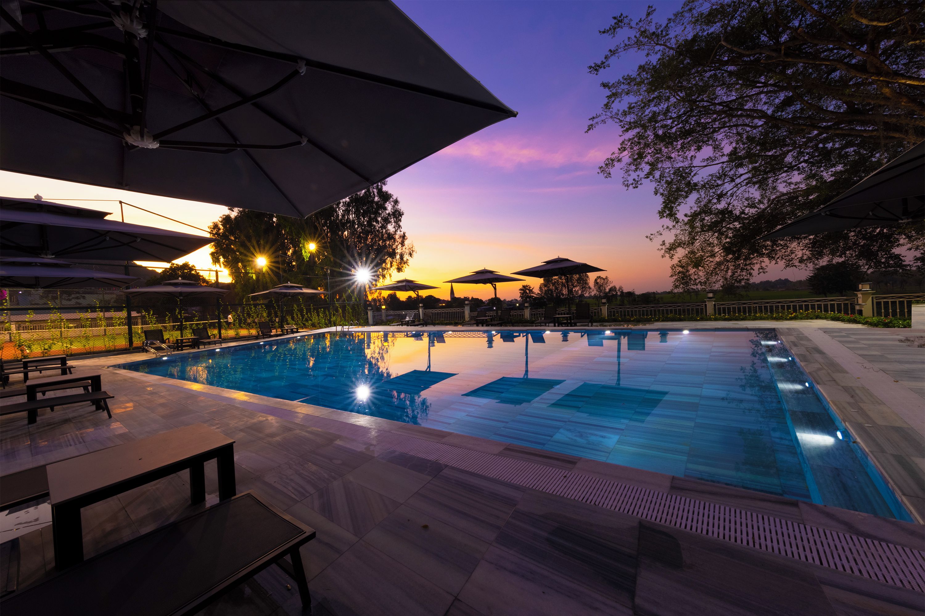 Una piscina con sillas de descanso y sombrillas, rodeada de árboles y un atardecer de fondo.