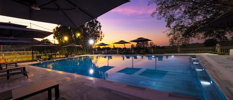 Una piscina con sillas de descanso y sombrillas, rodeada de árboles y un atardecer de fondo.