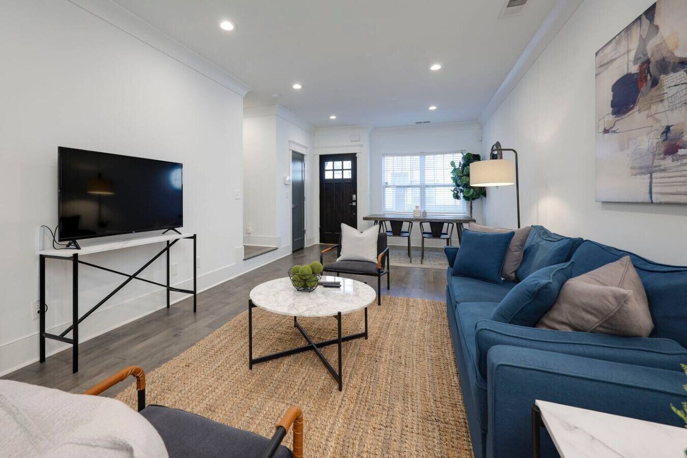 A modern living room with a flat-screen TV, a blue sofa, a coffee table, and a dining area in the background.
