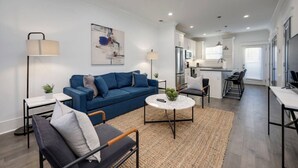 A modern living room with a blue sofa, a coffee table, and a dining area with a kitchen in the background.