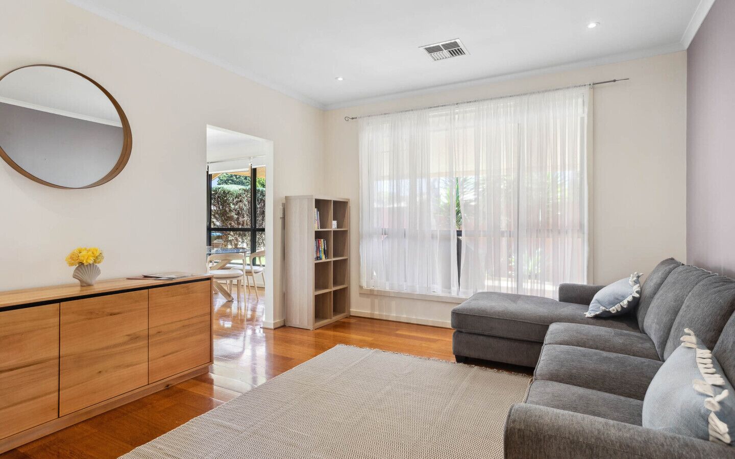 A modern living room with a grey sofa, wooden cabinet, and a large window with sheer curtains.