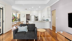 Una sala de estar moderna con un sofá gris, una mesita redonda y una zona de cocina con armarios y electrodomésticos blancos.