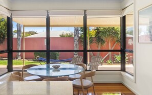 A sunlit room with a round glass table, four chairs, and a view of a red building and greenery outside. - Chiton Rocks Court! Mini Oasis by the Sea (Chiton)