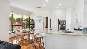 Una cocina moderna con gabinetes blancos, un refrigerador de acero inoxidable, un horno y un microondas. Hay una barra con taburetes y un ventanal con vistas a zonas verdes.