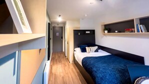 Quarto de hotel compacto com cama, prateleiras ao lado, uma escrivaninha pequena e vista para o corredor.