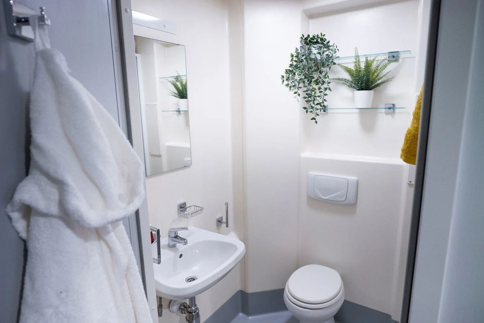 Un baño pequeño y moderno con lavamanos, espejo y retrete blancos. Hay dos plantas en macetas sobre estantes encima del inodoro.