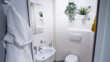 Un baño pequeño y moderno con lavamanos, espejo y retrete blancos. Hay dos plantas en macetas sobre estantes encima del inodoro.