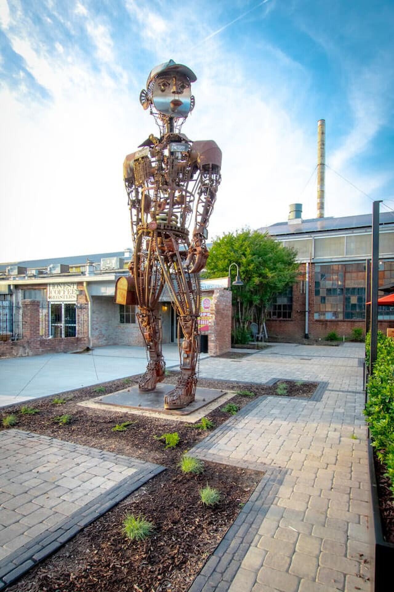 A large metal sculpture of a human figure in an outdoor plaza with brick pathways and greenery.