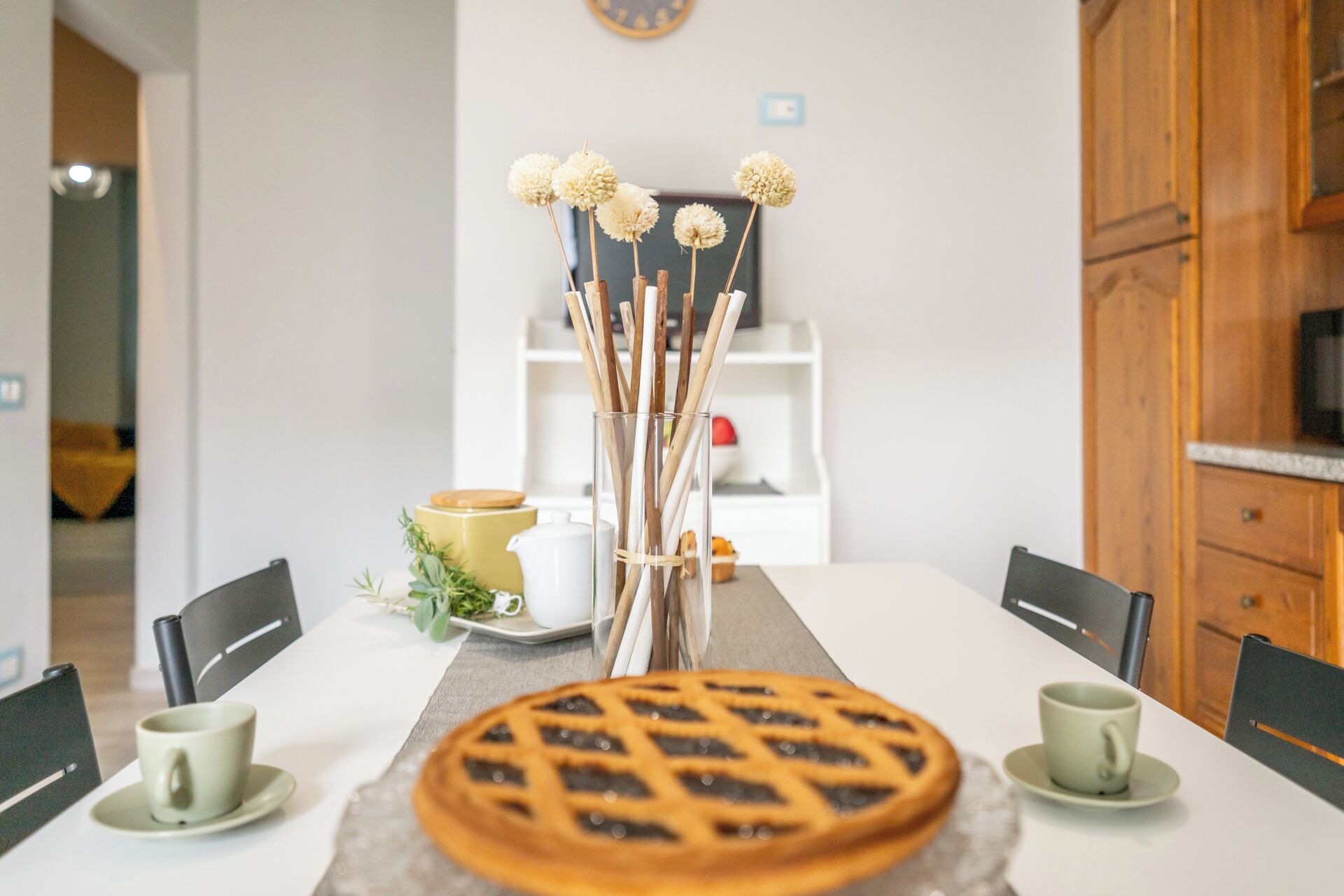 A dining table with a pie, two cups, and a vase with dried flowers.