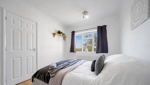 A bedroom with a bed, a window with curtains, a door, and a wall-mounted shelf with plants.
