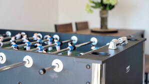 A foosball table with metal rods and figures, a dark-colored table, and a vase with flowers in the background.