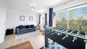 A modern living room with a foosball table, a grey sofa, and a large window offering a view of trees.