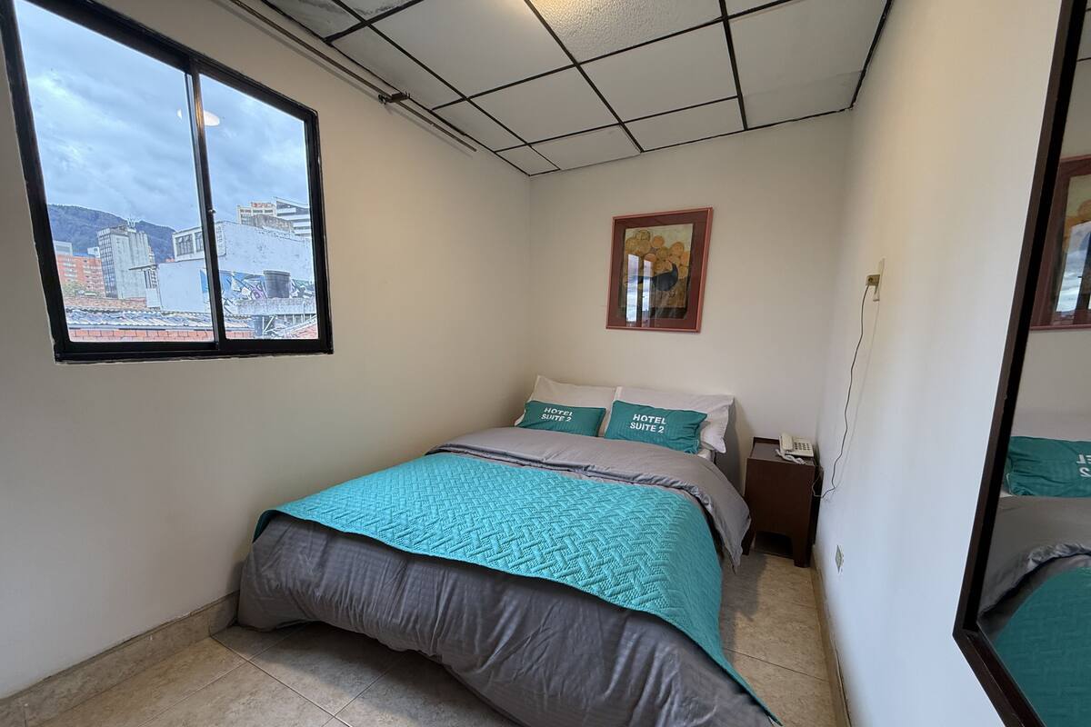 A bedroom with a bed, a window, a framed picture, and a bedside table with a phone.
