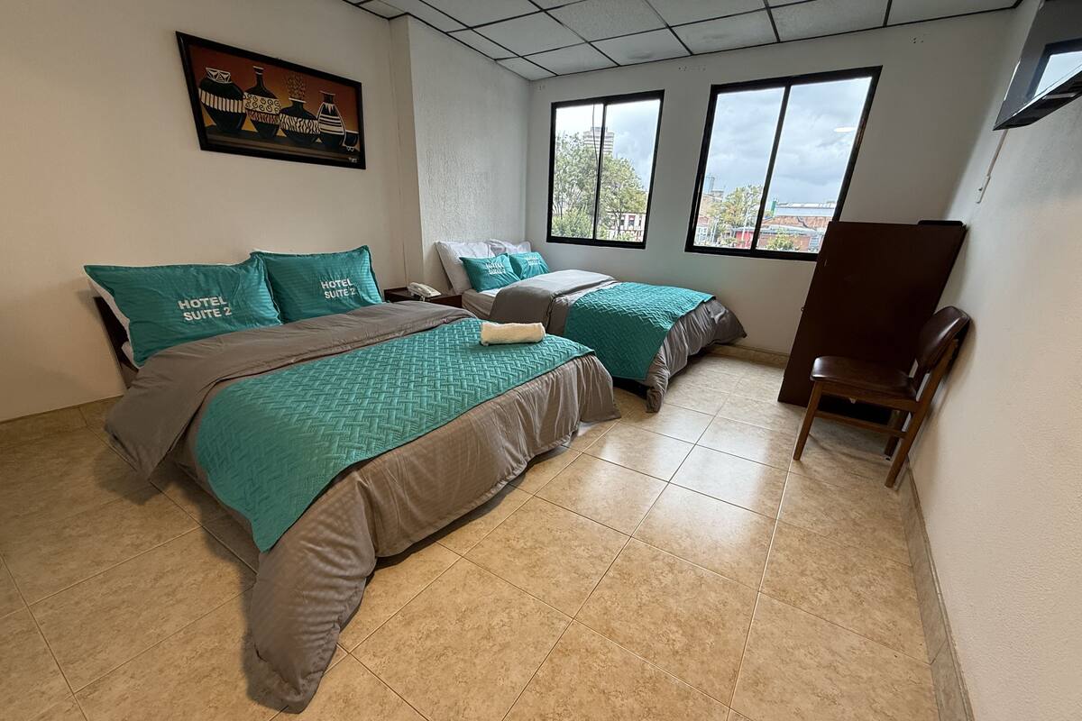 A hotel room with two beds, teal bedding, a painting on the wall, and a view through the windows.