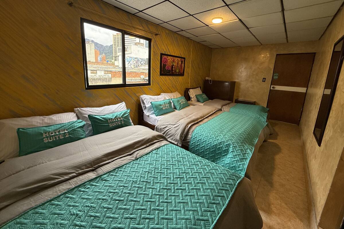 A hotel room with two beds, each with green bedding and pillows labeled "HOTEL SUITE 2". There is a window with a view of buildings and a door to the right.