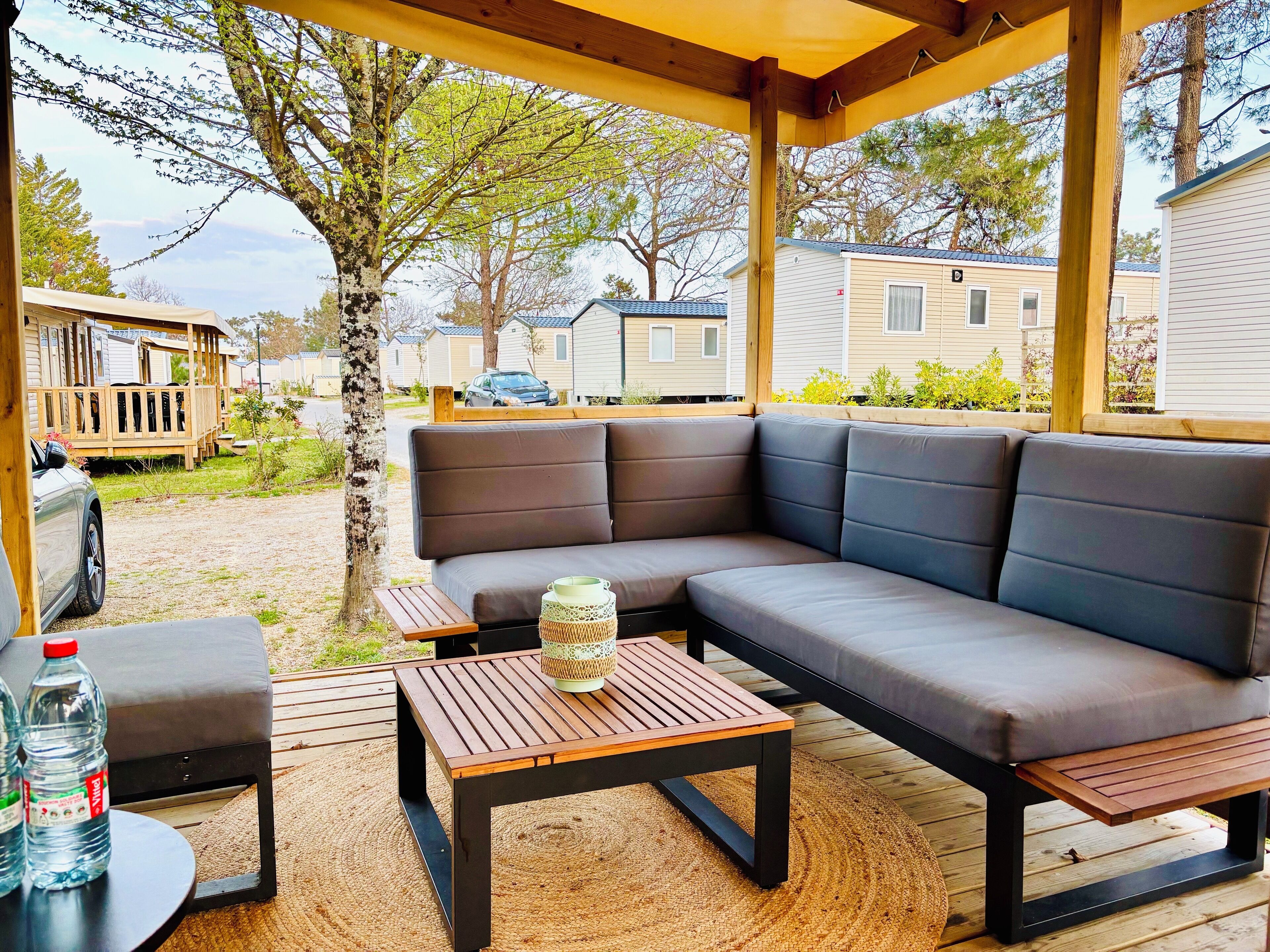 Uma área de estar coberta ao ar livre, com um sofá e duas mesas, cercada por um deck de madeira e uma pérgola.